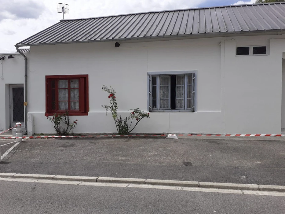 Maison blanche rénovée avec volets rouges et bleus, toiture métallique nervurée, rosiers plantés en façade